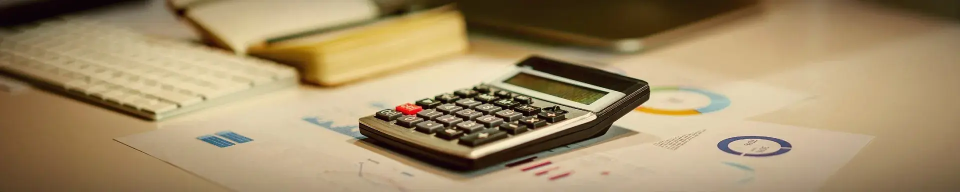 A desk calculator resting on printed financial reports and pie charts, with a keyboard and a bound notebook alongside, representing a sole trader weighing up their accounting options
