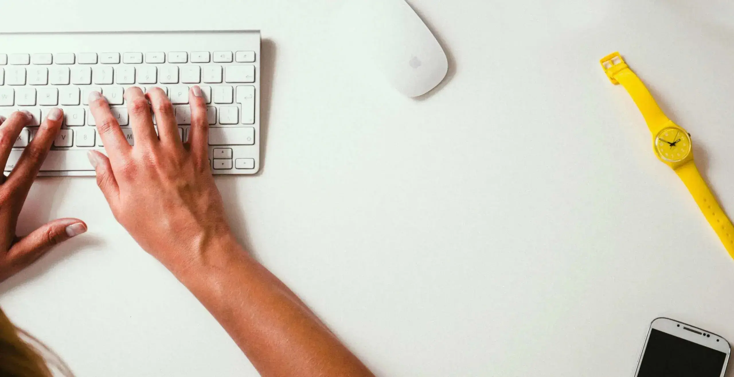 Person typing on a keyboard with a phone and watch on the desk