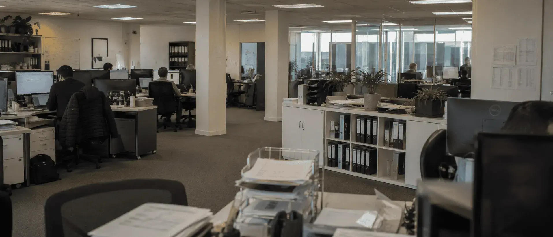 An open-plan office with staff working at desks in front of monitors, shelves of lever-arch binders along one wall and stacked paperwork trays in the foreground